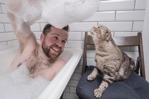Ein zufriedener Mann badet in einer Badewanne, er hält eine üppige Seifenlauge in der Hand, an der sich seine angebetete Katze interessiert. foto