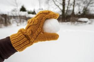 Eine Hand in einem gelben Fäustling hält einen Schneeball im Hof vor dem Hintergrund eines schneebedeckten Gemüsegartens. Winteraktives Spiel und Spaß. foto