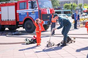 Ein Feuerwehrmann bringt einem kleinen Mädchen in einem aufdringlichen feuerfesten Anzug bei, mit Schläuchen herumzulaufen, um Poren zu löschen Weißrussland, Minsk, 08.08.2018 foto