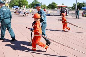 Ein Feuerwehrmann bringt einem kleinen Mädchen in einem aufdringlichen feuerfesten Anzug bei, mit Schläuchen herumzulaufen, um Poren zu löschen Weißrussland, Minsk, 08.08.2018 foto