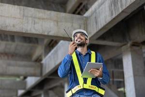 Der kaukasische Ingenieur mit Funk-Walkie-Talkie in voller Sicherheitsausrüstung inspiziert die Gebäudestruktur zur Untersuchung des Spezifikations- und Qualitätskontrollkonzepts foto