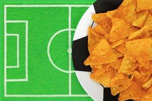 Nacho-Chips aus mexikanischem Mais auf einem großen Teller, der wie ein Fußball bemalt ist, und eine Geschirrmatte wie ein Fußballfeld aus grünem Filz mit Platz für Text. vorlage des fußballpartytisches. draufsicht, kopierraum, modell. foto