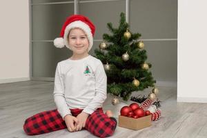 mädchen im karierten pyjama und eine weihnachtsmütze sitzt neben dem weihnachtsbaum und lächelt, weihnachten foto