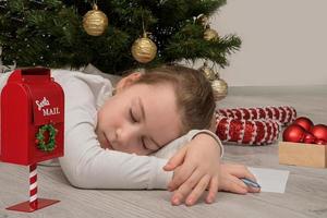 Das kleine Mädchen schlief unter dem Weihnachtsbaum ein, während es einen Brief an den Weihnachtsmann schrieb foto