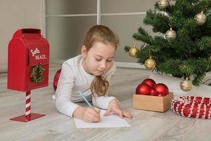 mädchen im pyjama schreibt einen brief an den weihnachtsmann in der nähe des weihnachtsbaums, weihnachten, weihnachtsgeschenke foto