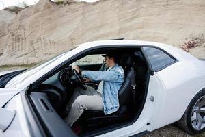 Schöner Mann in Jeansjacke und Mütze sitzt an seinem weißen Muscle-Car. foto