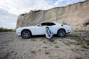 Gutaussehender Mann in Jeansjacke und Mütze, der in der Karriere neben seinem weißen Muscle-Car sitzt. foto