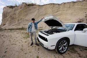 Schöner Mann in Jeansjacke und Mütze steht neben seinem weißen Muscle-Car mit aufklappbarer Motorhaube, Motor prüfen. foto