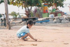 Asiatisches süßes kleines Mädchen, das Sandburgen spielt oder baut oder mit Sand am tropischen Strand gräbt. Kinder mit schönem Meer, Sand und blauem Himmel. glückliche kinder im urlaub am meer, die am strand laufen. foto
