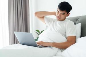 asiatischer mann in weißem t-shirt, der mit einem laptop-computer mit nackenschmerzen auf dem bett liegt. foto