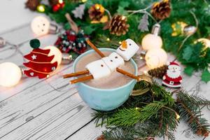 große Tasse mit heißem Cappuccino mit geschmolzenem Marshmallow-Schneemann auf einem Hintergrund von Bokeh-Weihnachtsgirlanden und Weihnachtsbaum. foto