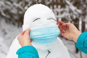 schneemann in einer medizinischen maske - eine neue realität, schutz vor krankheiten, infektionen, leben in der covid-epidemie. Frauenhände setzen eine Schneemannmaske auf. Outdoor-Aktivitäten für die Winterfamilie foto