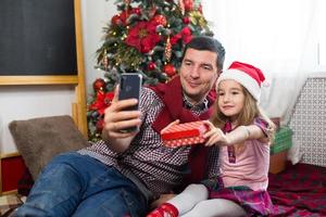 vater und tochter in der nähe des weihnachtsbaums mit einem smartphone machen ein selfie, kommunizieren über videoverbindung. weihnachtsgrüße, eine geschenkbox, ein mädchen in einer weihnachtsmütze winkt hallo. neujahr, feiertagsdekor foto