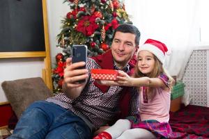 vater und tochter in der nähe des weihnachtsbaums mit einem smartphone machen ein selfie, kommunizieren über videoverbindung. weihnachtsgrüße, eine geschenkbox, ein mädchen in einer weihnachtsmütze winkt hallo. neujahr, feiertagsdekor foto