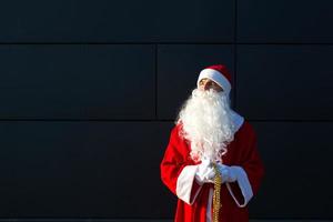 ein junger mann, der als weihnachtsmann auf der straße verkleidet ist und an einer grauen modernen wand steht. weißer Bart, ein roter Anzug mit Hut. weihnachten, neujahr. Russischer Großvater Frost. grauer moderner hintergrund, kopierräume foto