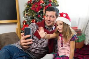 vater und tochter in der nähe des weihnachtsbaums mit einem smartphone machen ein selfie, kommunizieren über videoverbindung. weihnachtsgrüße, eine geschenkbox, ein mädchen in einer weihnachtsmütze winkt hallo. neujahr, feiertagsdekor foto