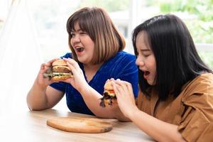 hungrige übergewichtige frau, die hamburger auf einer holzplatte hält, während der arbeit von zu hause, gewichtsproblem. Konzept des Binge-Eating-Störungsbettes. foto