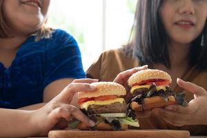 hungrige übergewichtige frau, die hamburger auf einer holzplatte hält, während der arbeit von zu hause, gewichtsproblem. Konzept des Binge-Eating-Störungsbettes foto