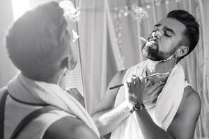 Mann, der in den Spiegel schaut, eine Zigarette raucht und seinen Bart mit einem Rasiermesser rasiert foto