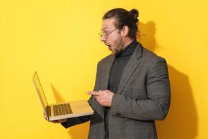 Überraschter Mann mit Brille, der auf Laptop-Bildschirm auf gelbem Hintergrund zeigt foto