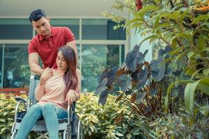 asiatische Frau im Rollstuhl und unglücklich und schmerzhaft. Ein Mann steht hinter dem Rollstuhl und macht seiner Frau Mut, deren Füße sich durch einen Unfall am Bein verletzt haben. Konzept der Fürsorge und Unterstützung