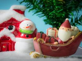 glücklicher weihnachtsmann mit geschenkbox auf dem schneeschlitten, der zum haus geht. in der nähe des hauses haben schneemann und weihnachtsbaum. weihnachtsmann und haus im schnee. weihnachts- und frohes neues jahrkonzept. foto