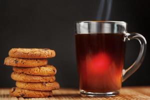 Ein Haufen Haferkekse mit Schokoladenstückchen und eine Tasse duftenden schwarzen heißen Tees auf einem Bambussubstrat auf dunklem Hintergrund. handgemachte Kekse für ein gesundes Frühstück foto