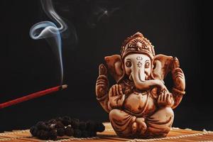 Hindu-Gott Ganesh auf schwarzem Hintergrund. Rudraksha-Statue und Rosenkranz auf einem Holztisch mit rotem Räucherstäbchen und Weihrauchrauch foto