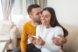 Fröhliches Paar, das zu Hause die Ergebnisse eines Schwangerschaftstests herausfindet. glückliches Paar, das sich einen Schwangerschaftstest ansieht. Frau, die ihren Mann mit einem positiven Schwangerschaftstest überrascht, scheint er einigermaßen zufrieden zu sein foto