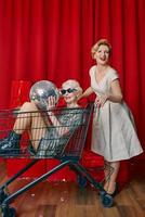 reife Frau rollt stilvolle Seniorin mit Sonnenbrille und silbernem Kleid im Einkaufswagen auf der Party. party, disko, feier, seniorenkonzept foto