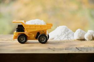 Ein kleiner gelber Spielzeuglastwagen ist mit einem Stein aus weißem Salz neben einem Salzhaufen beladen. ein Auto auf einer Holzoberfläche vor dem Hintergrund eines herbstlichen Waldes. Gewinnung und Transport von Salz foto
