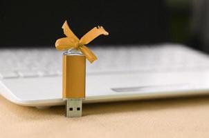 Eine orangefarbene USB-Flash-Speicherkarte mit einer Schleife liegt auf einer Decke aus weichem und pelzigem, hellorangefarbenem Fleece-Stoff neben einem weißen Laptop. klassisches weibliches Geschenkdesign für eine Speicherkarte foto