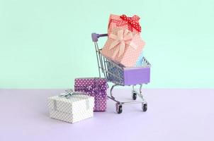 kleine geschenkboxen in verschiedenen farben mit bändern liegen im einkaufswagen auf violettem und blauem pastellhintergrund foto