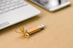 Eine orangefarbene USB-Flash-Speicherkarte mit blauer Schleife liegt auf einer Decke aus weichem, pelzigem, hellorangefarbenem Fleece-Stoff neben einem weißen Laptop und einem Smartphone. klassisches weibliches Geschenkdesign für eine Speicherkarte foto