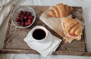 leckeres frühstück mit frischem croissant, kaffee, kirschen auf einem holztablett. herzhaftes Croissant mit Tomate und Käse. Espresso auf einem Frühstückstablett foto