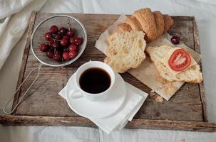 leckeres frühstück mit frischem croissant, kaffee, kirschen auf einem holztablett. herzhaftes Croissant mit Tomate und Käse. Espresso auf einem Frühstückstablett foto