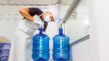 mann arbeiter in arbeitskleidung und mit einer schutzmaske im gesicht, die in einer trinkwasserfabrik arbeitet, die blaue wassergallonen vor dem versand überprüft. foto