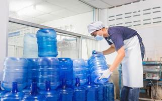 mann arbeiter in arbeitskleidung und mit einer schutzmaske im gesicht, die in einer trinkwasserfabrik arbeitet, die blaue wassergallonen vor dem versand überprüft. foto