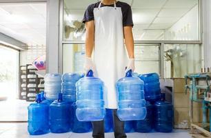 mann arbeiter in arbeitskleidung und mit einer schutzmaske im gesicht, die in einer trinkwasserfabrik arbeitet, die blaue wassergallonen vor dem versand überprüft. foto