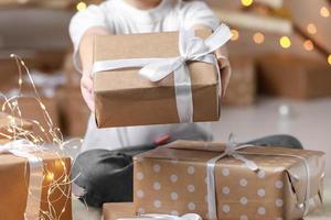 einfaches geschenk in kraftpapierverpackungen, die mit einem weißen band in den händen eines kinderjungen im hemd gebunden sind. Baby, das Geschenkbox auf Weihnachtshintergrund hält. konzept von neujahr, muttertag, valentinstag foto