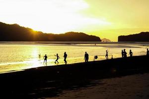 Silhouettengruppe von Menschen, die im Sonnenuntergang Fußball am Strand spielen, mit Meereslandschaft und Bergen im Hintergrund foto