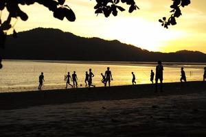 Silhouettengruppe von Menschen, die im Sonnenuntergang Fußball am Strand spielen, mit Meereslandschaft und Bergen im Hintergrund foto