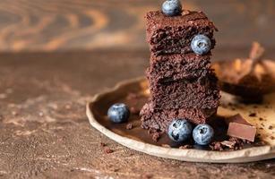 Schokoladenbrownie-Quadrate, verziert mit Heidelbeeren und Stücken dunkler Schokolade und Kakaopulver auf handwerklicher Keramikplatte, brauner Hintergrund. foto