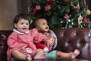 Zwei verschiedene Babyfreunde lächeln glücklich, während sie mit Geschenken auf der Couch spielen, die sich in Weihnachtskostümen und Weihnachtsmützen mit Weihnachtsbaum auf dem Rücken für das Festkonzept der Saison kleiden foto
