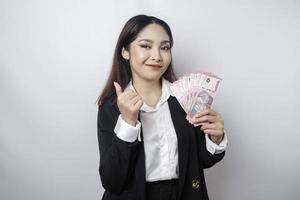 Aufgeregte asiatische Geschäftsfrau im schwarzen Anzug gibt Daumen hoch Handgeste der Zustimmung und hält Geld in indonesischer Rupiah, isoliert durch weißen Hintergrund foto