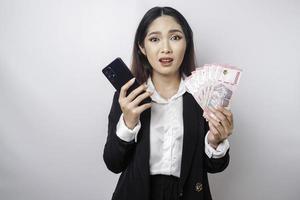 eine verwirrte junge geschäftsfrau trägt einen schwarzen anzug und hält ihr telefon und geld in indonesischer rupiah, isoliert durch einen weißen hintergrund foto