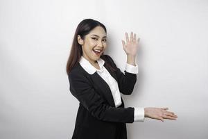Aufgeregte asiatische Geschäftsfrau im schwarzen Anzug, die auf den Kopierbereich neben ihr zeigt, isoliert durch weißen Hintergrund foto
