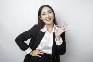Aufgeregte asiatische Geschäftsfrau, die einen schwarzen Anzug trägt und eine ok-Handgeste gibt, die von einem weißen Hintergrund isoliert wird foto