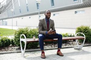 Afroamerikaner-Geschäftsmann arbeitet in seinem Smartphone, das draußen auf der Bank sitzt