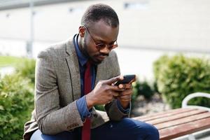 Afroamerikaner-Geschäftsmann arbeitet in seinem Smartphone, das draußen auf der Bank sitzt foto